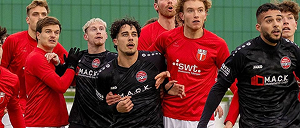Eine Gruppe männlicher Sportler in roten und schwarzen Trikots steht eng beieinander auf dem Spielfeld und blickt konzentriert in eine Richtung, vermutlich während einer Spielunterbrechung.