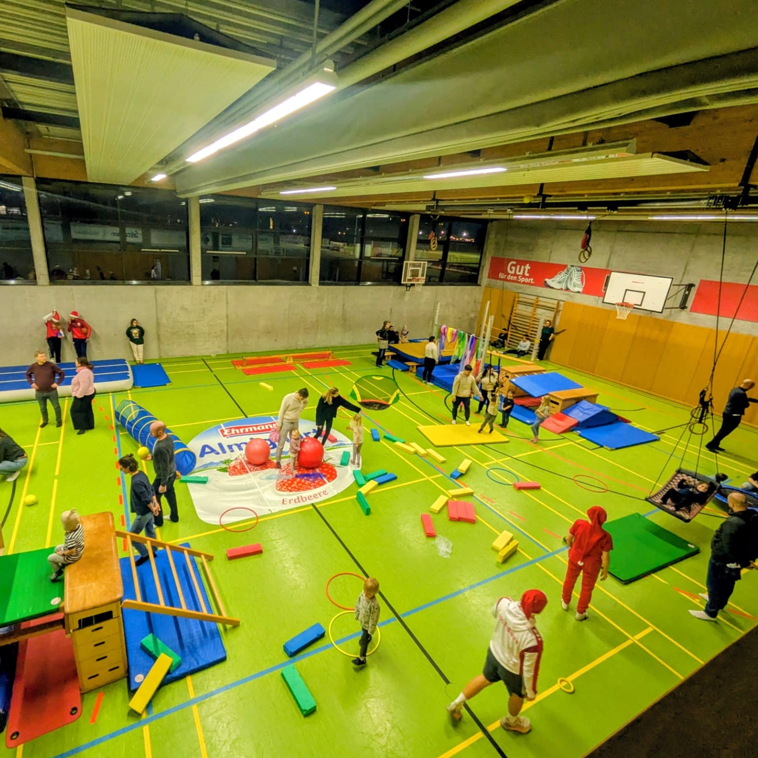 Kinder und Erwachsene in einer Turnhalle mit bunten Sportgeräten, Matten und Kletterelementen beim Spielen und Bewegen