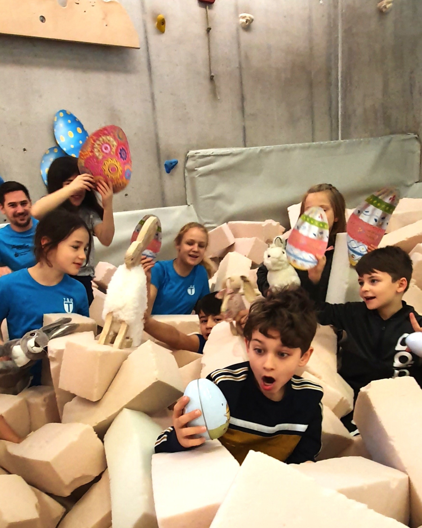 Kinder und Erwachsene feiern Ostern in einer Kletterhalle, umgeben von Schaumstoffblöcken, bunten Eiern und Hasen.