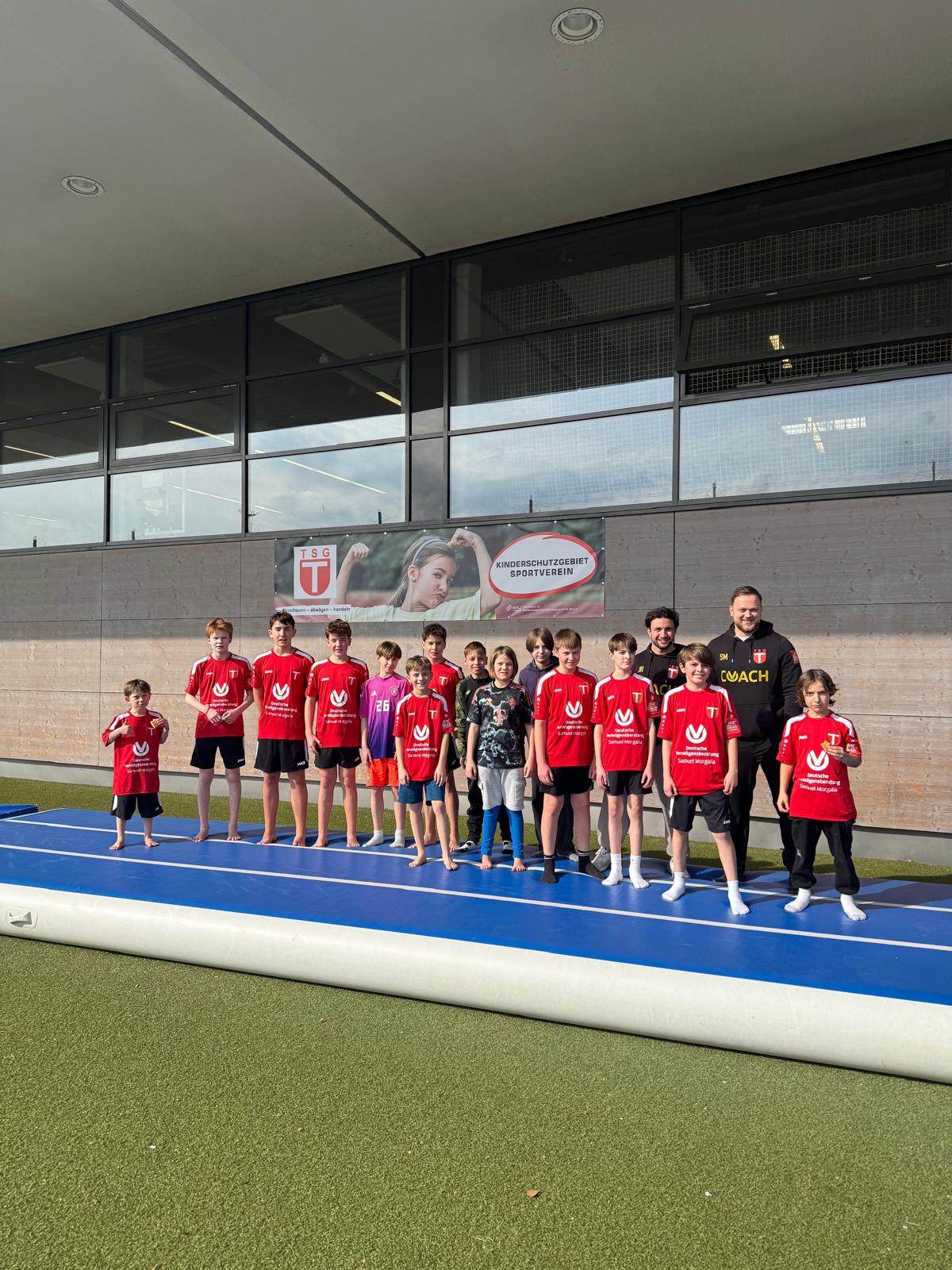 Jugendliche Fußballmannschaft und zwei Trainer stehen barfuß auf einer blauen Matte vor einer Hallenwand mit Vereinsbanner.