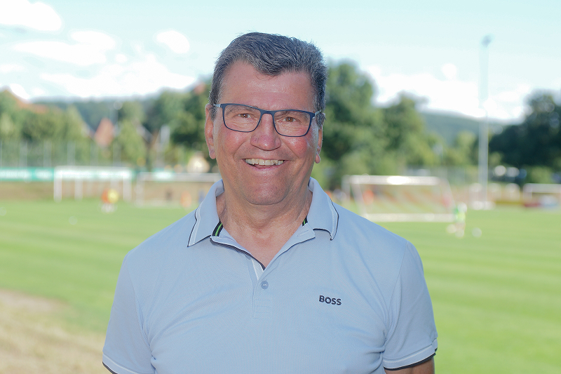 Ein lächelnder Mann mit Brille steht vor einem Fußballfeld, im Hintergrund sind Tore und Spieler auf dem Platz zu sehen.