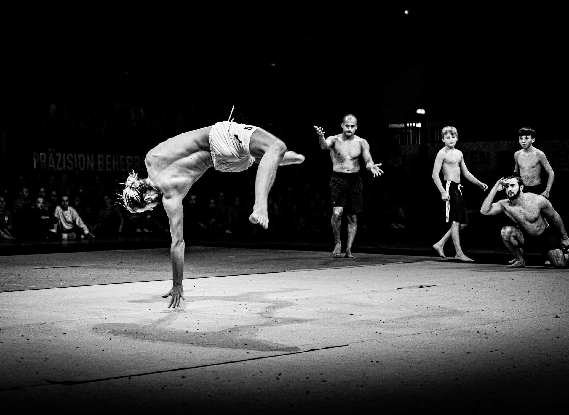 Ein Akrobat hält sich bei einer Bühnenshow in einer Kraftfigur im einarmigen Handstand auf dem Boden. Weitere Zuschauer stehen im Hintergrund.