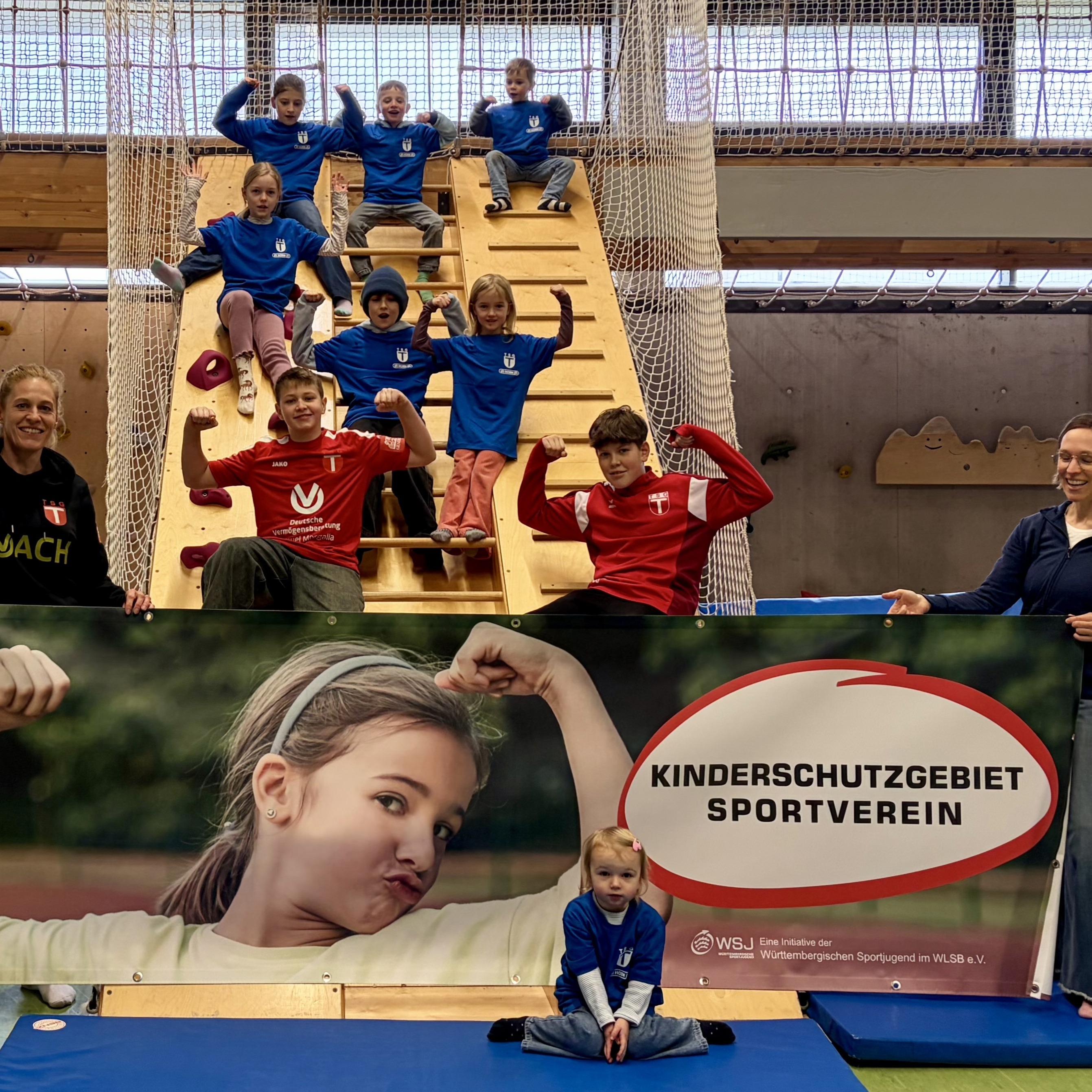 Kinder auf einer Kletteranlage in der Sporthalle, teils mit erhobenen Armen, vor einem Banner mit der Aufschrift „Kinderschutzgebiet Sportverein“.