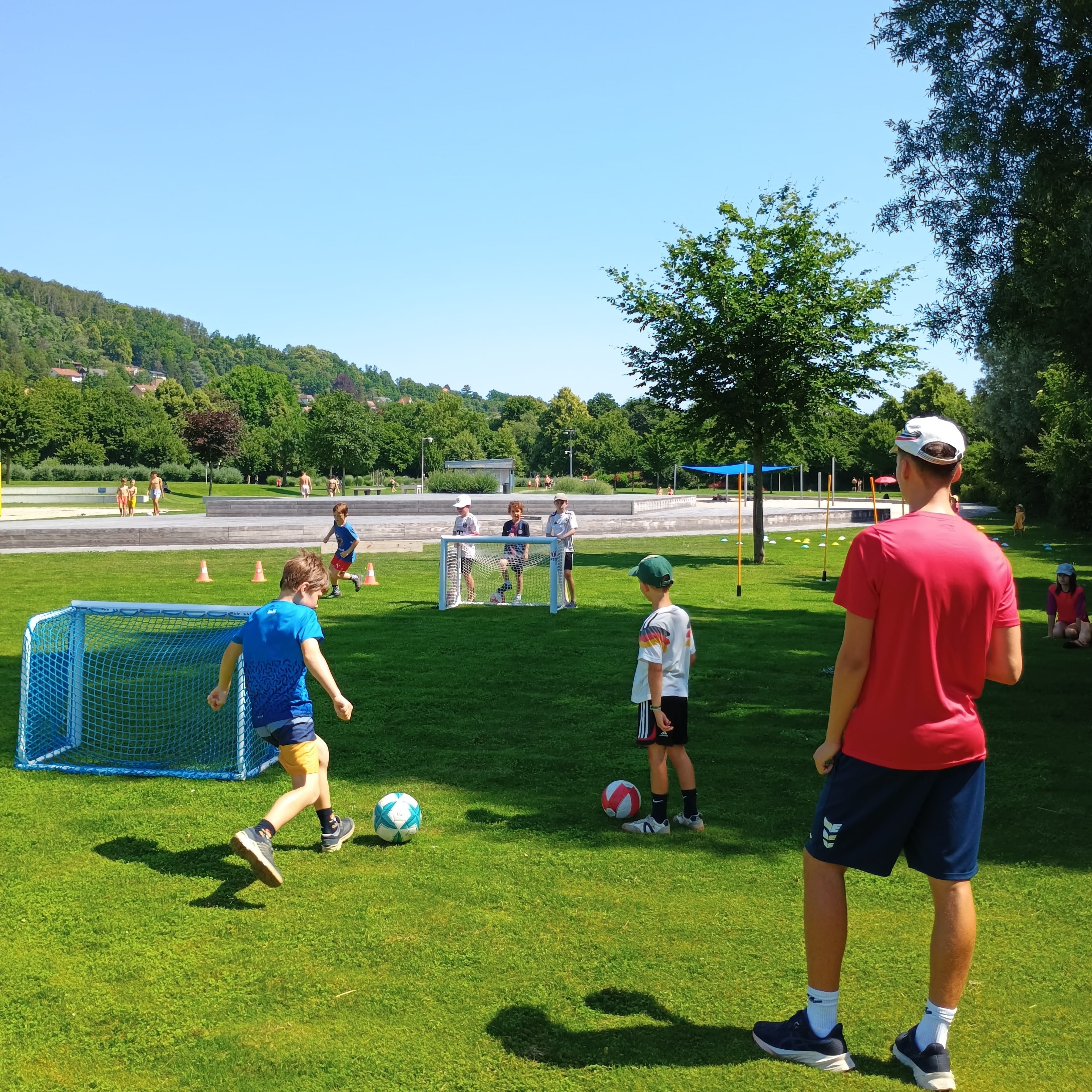 Kinder spielen Fussball auf sonniger Rasenflaeche mit kleinen Toren. Ein Erwachsener betreut die Gruppe, im Hintergrund Baeume und Huegel.