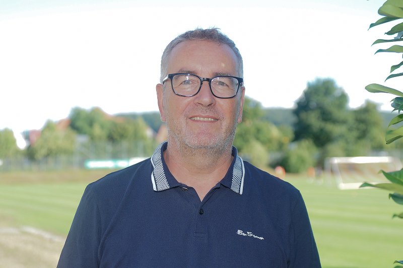 Ein Mann mit Brille und Polohemd steht im Freien vor einem Fußballfeld, umgeben von grüner Vegetation und Bäumen.