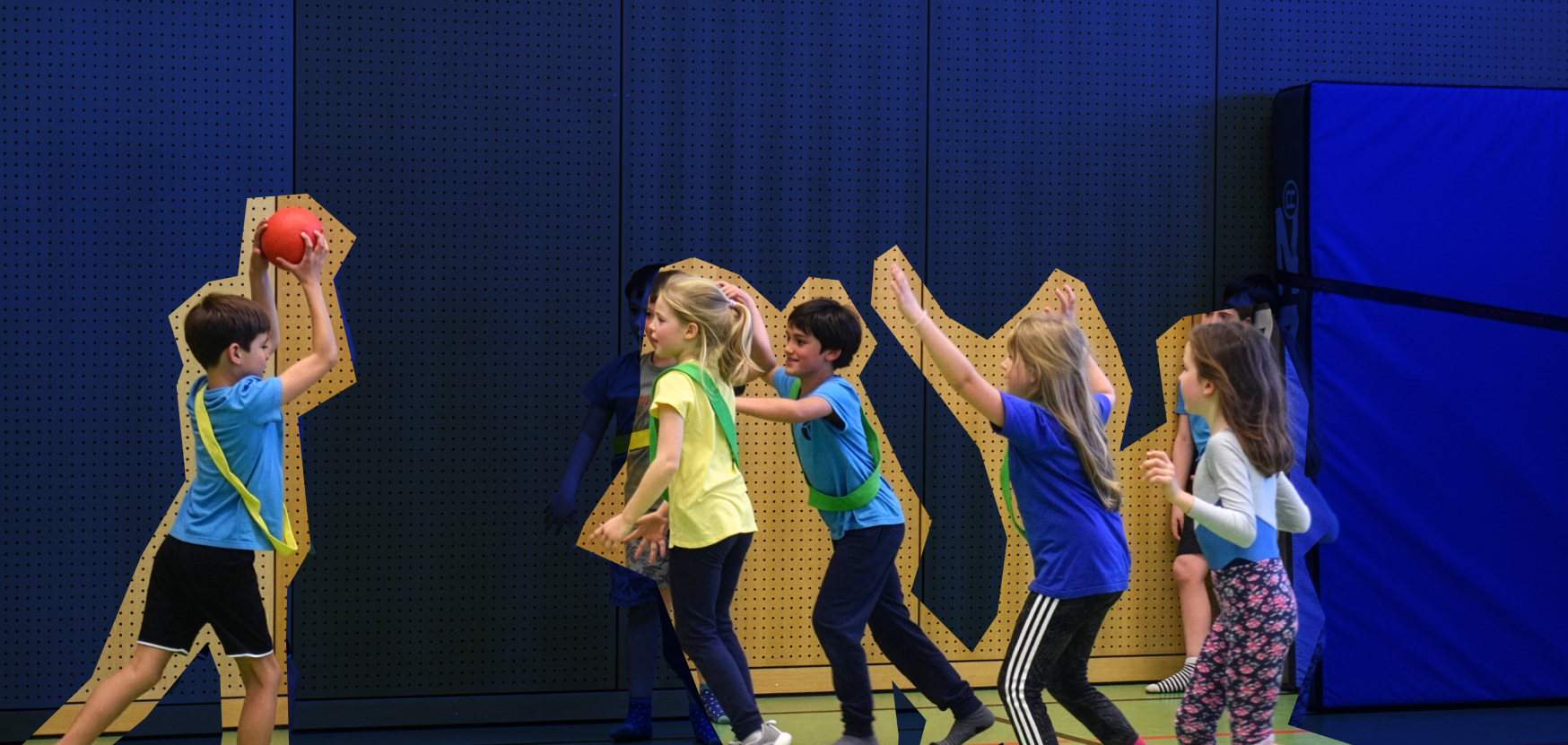Kinder spielen in einer Turnhalle Handball, während sie sich mit bunten Bändern bewegen. Im Hintergrund ist eine gepolsterte Wand zu sehen.