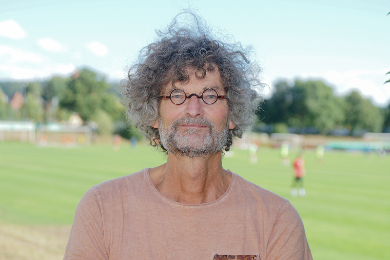 Mann mit lockigem, grauen Haar und Brille trägt ein helles T-Shirt, im Hintergrund ist ein Fußballfeld mit Spielern zu sehen.