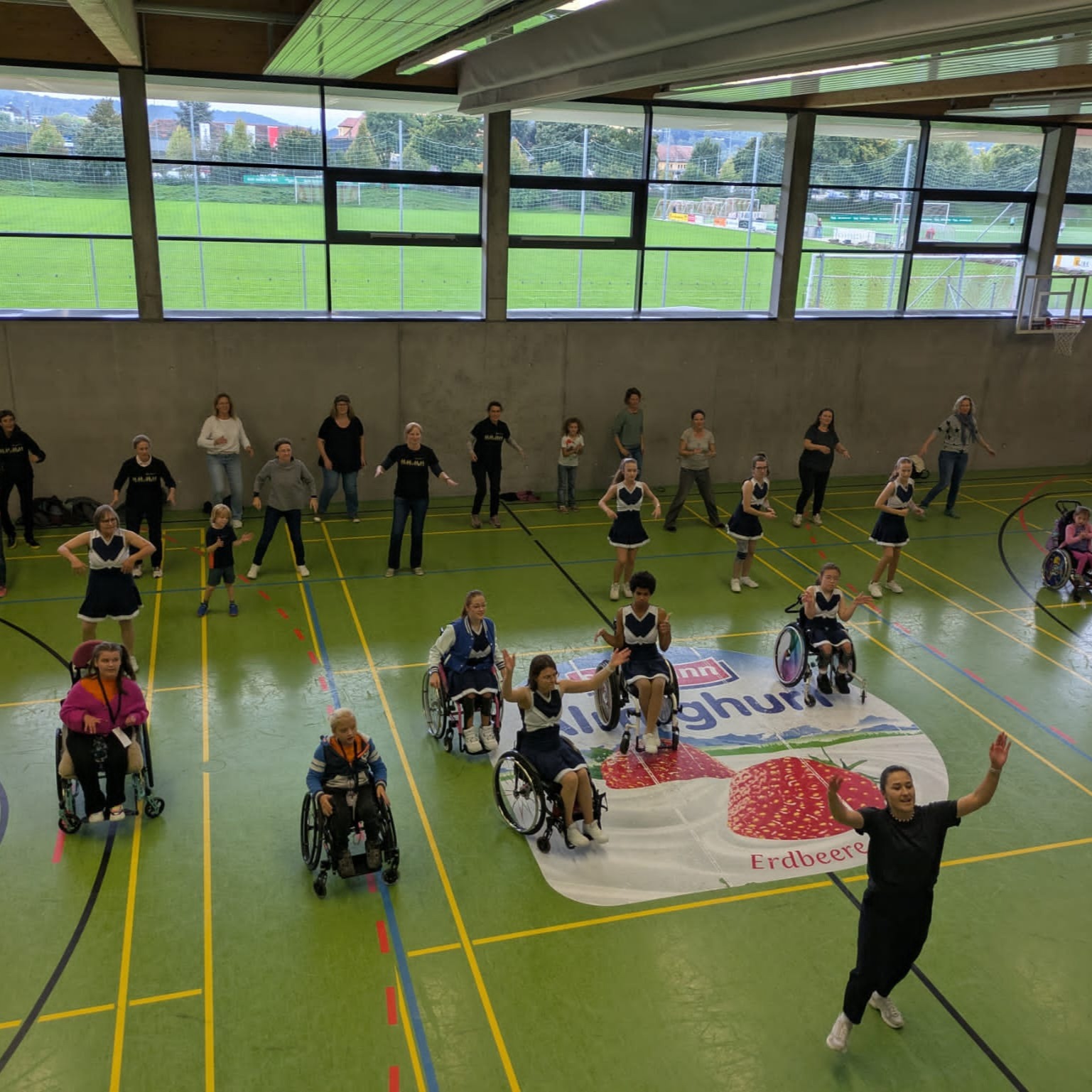 Menschen mit und ohne Rollstuhl üben gemeinsam eine Tanzchoreografie in einer Sporthalle; im Hintergrund ein Fußballplatz.
