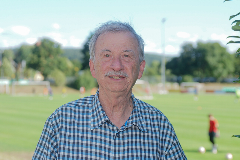 Älterer Mann im karierten Hemd vor einem Fußballplatz mit Spielern im Hintergrund.