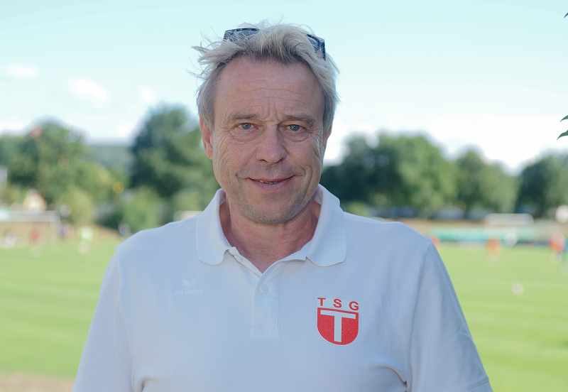 Mann im weißen Poloshirt mit TSG-Logo vor einem Sportplatz im Freien