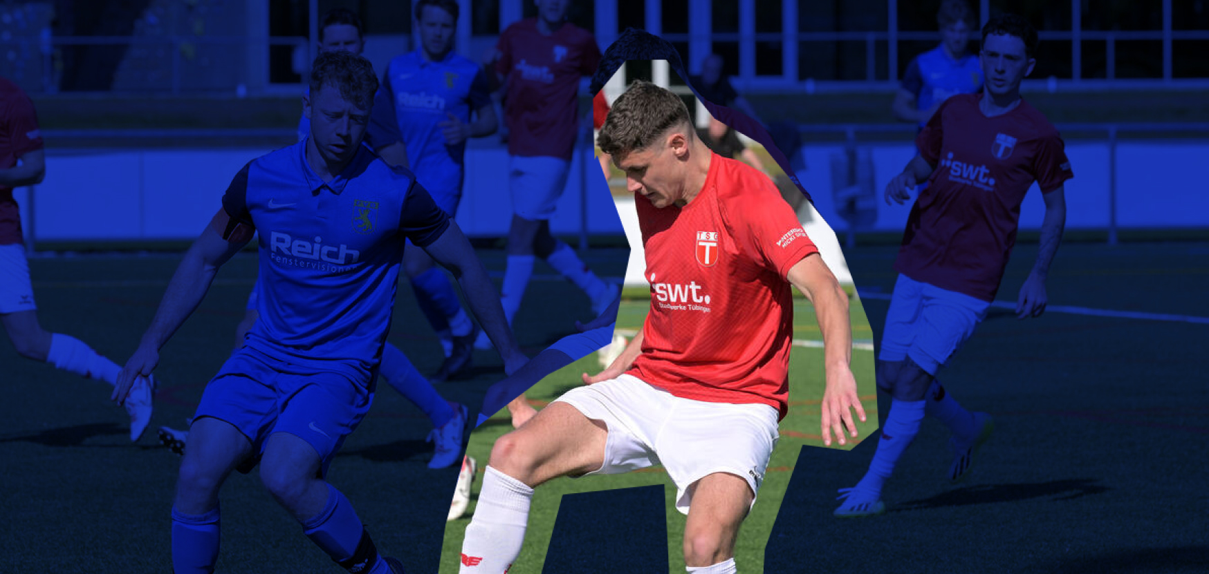 Zwei Fußballspieler auf dem Feld, einer trägt ein rotes Trikot und ballführt, der andere in blau verfolgt ihn. Hintergrund zeigt weitere Spieler und ein Sportg…