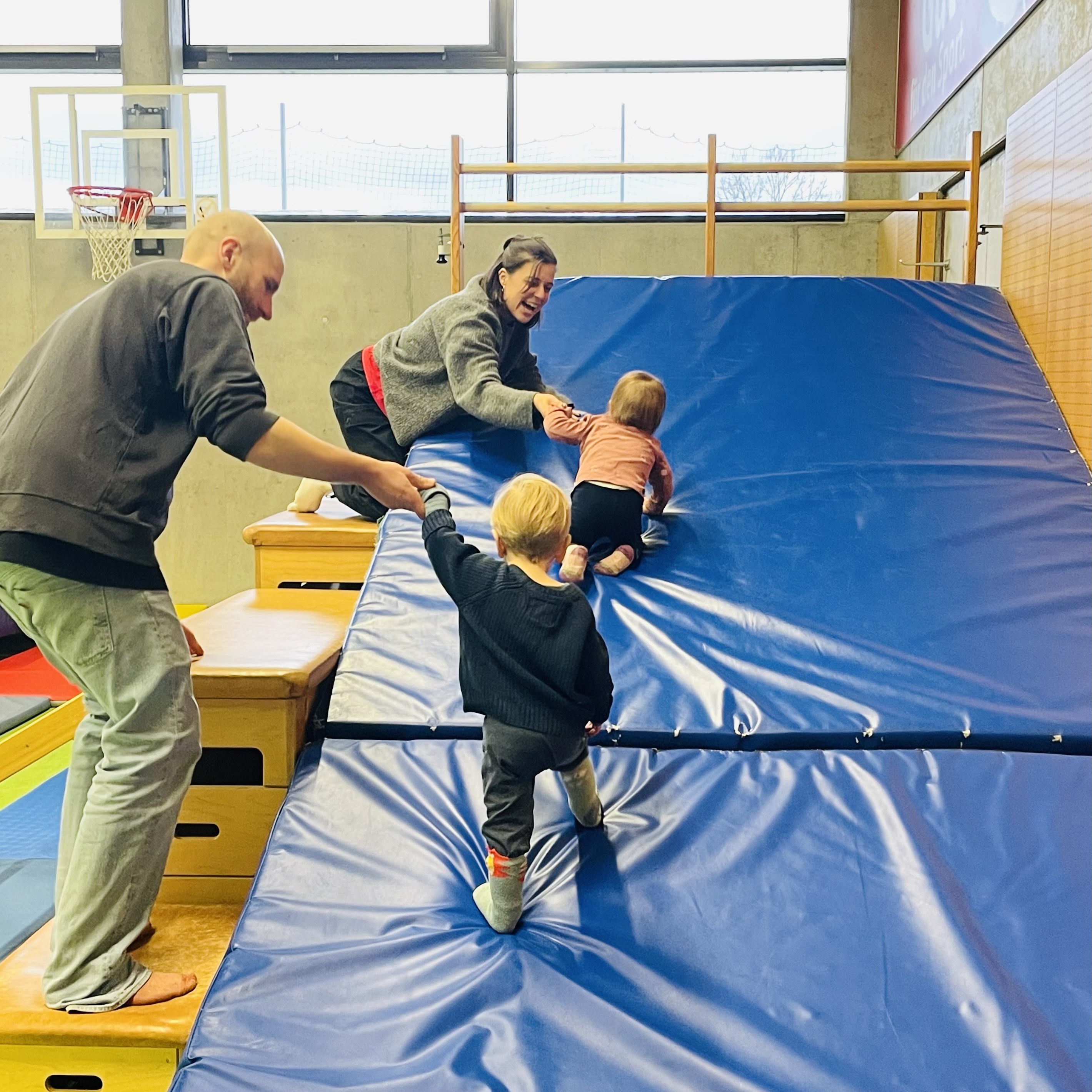 Erwachsene begleiten drei Kleinkinder auf einer blauen Turnmatte in einer Sporthalle.