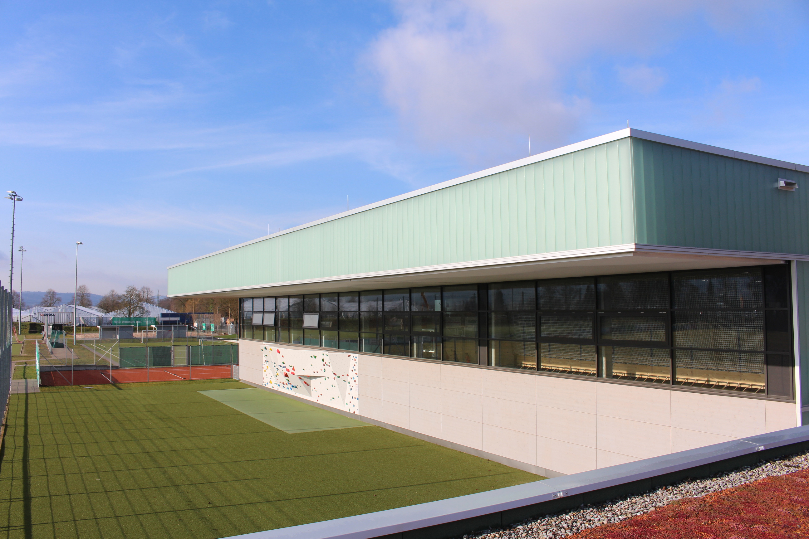 Außenansicht des modernen Sportzentrum-Gebäudes mit einer markanten Dachkonstruktion, großen Glasfronten und einer davor gelagerten Grünfläche.