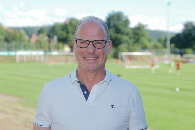 Lächelnder Mann mit Brille und weißem Poloshirt vor einem Fußballplatz mit Toren und Spielern im Hintergrund
