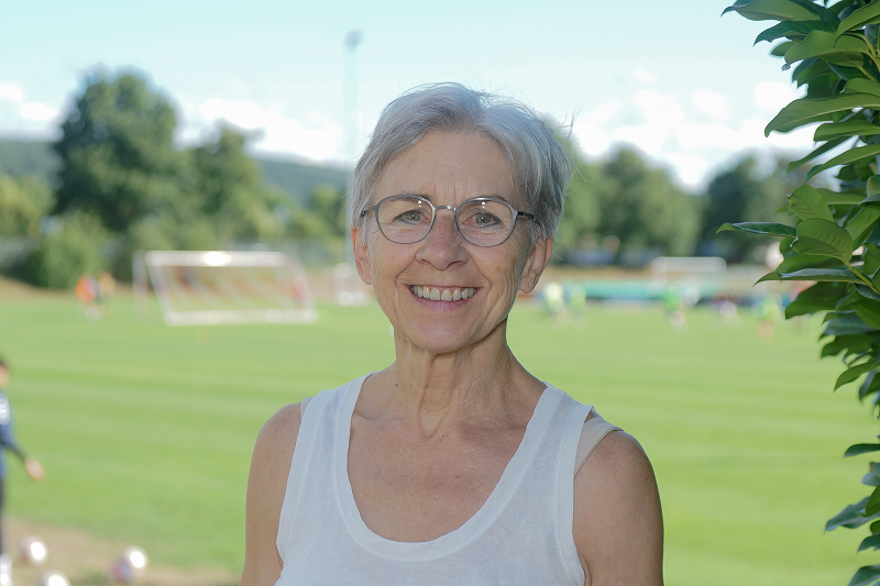 Ältere Frau mit Brille und weißem Top vor einem Fußballplatz im Freien