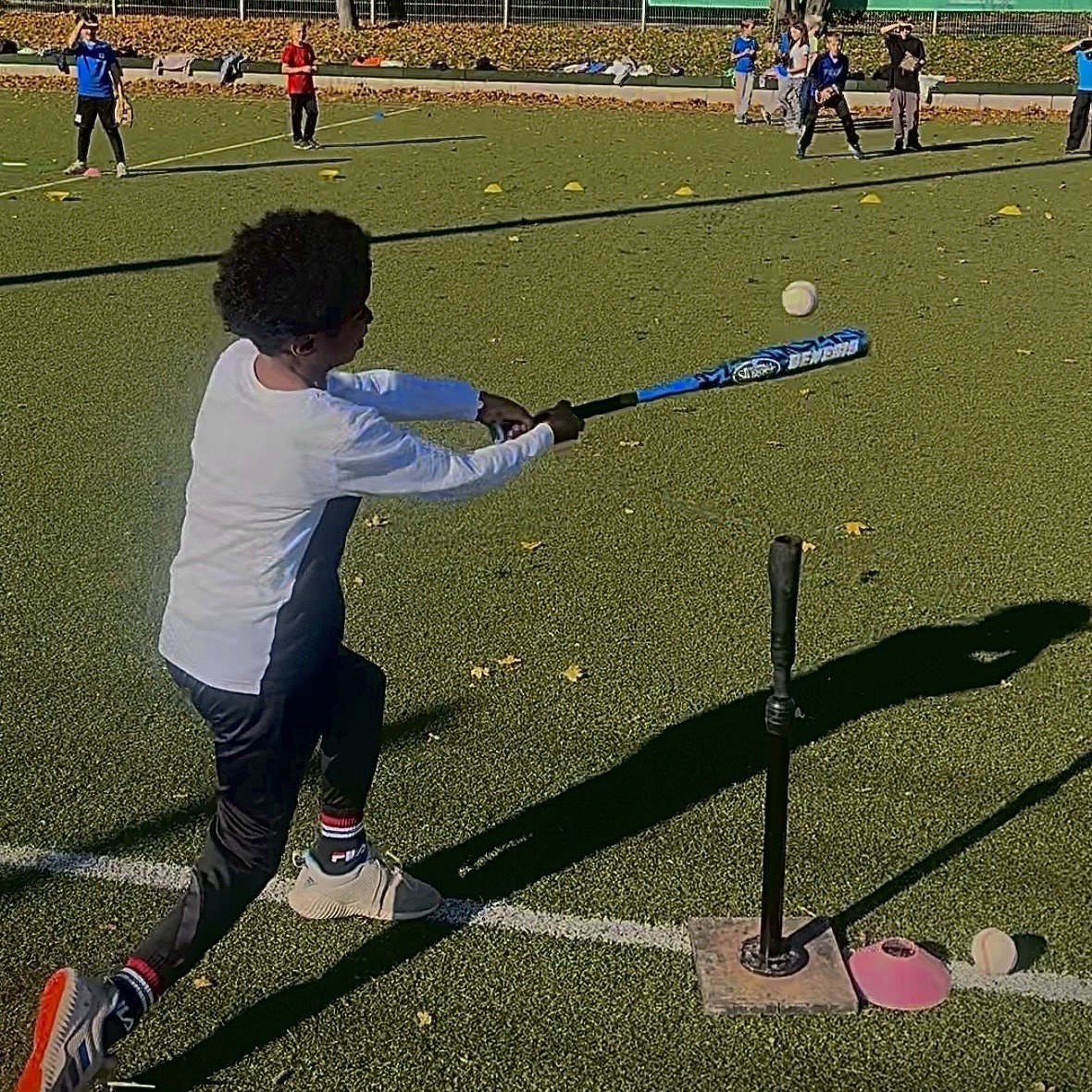 Junger Mensch schlägt einen Baseball mit blauem Schläger vom Batting-Tee auf grünem Sportplatz. Andere Kinder trainieren im Hintergrund.