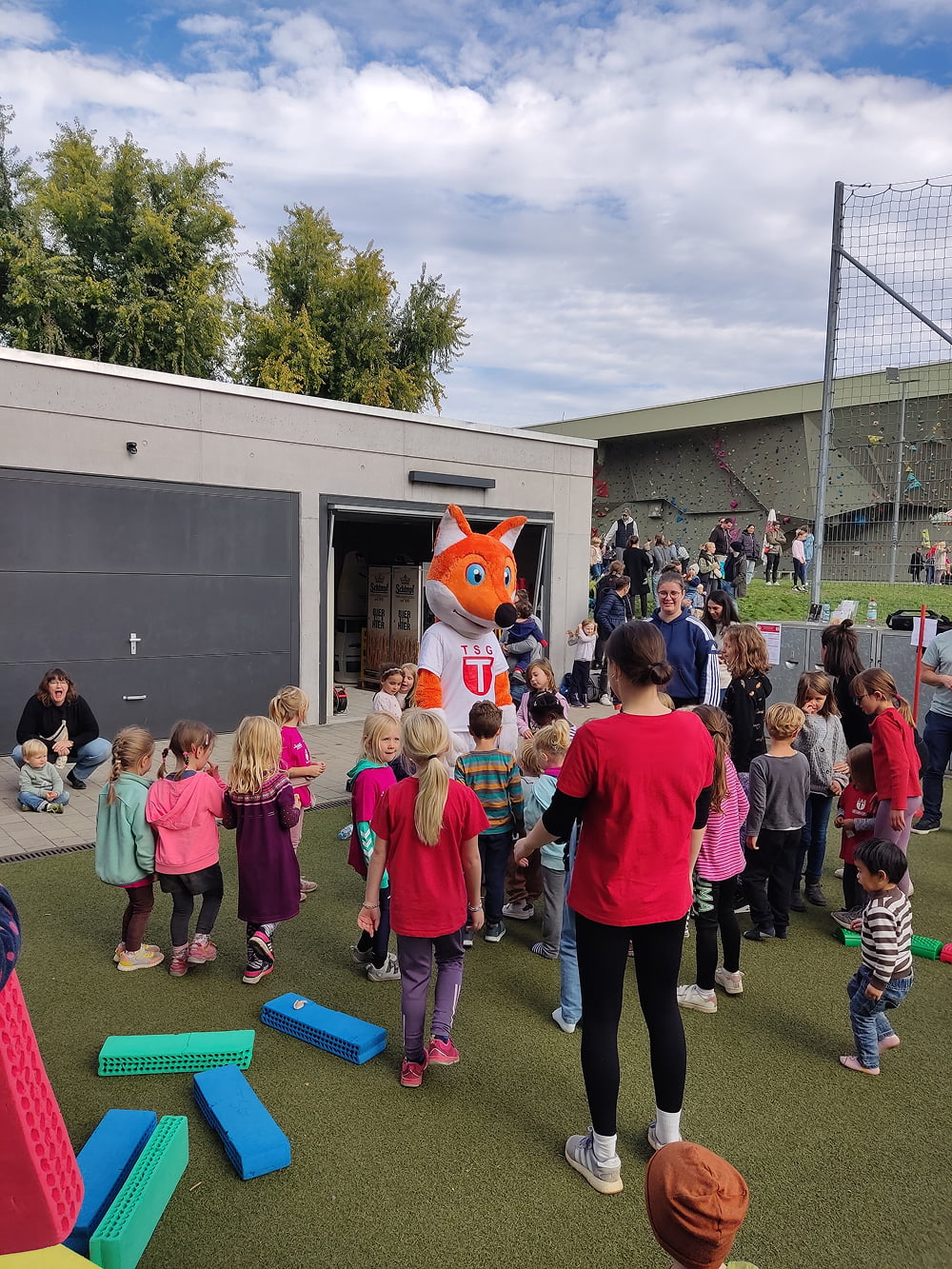 Kindergruppe bei einer Veranstaltung mit Fuchs-Maskottchen auf einem Sportplatz, daneben Zuschauer und eine Kletterwand.