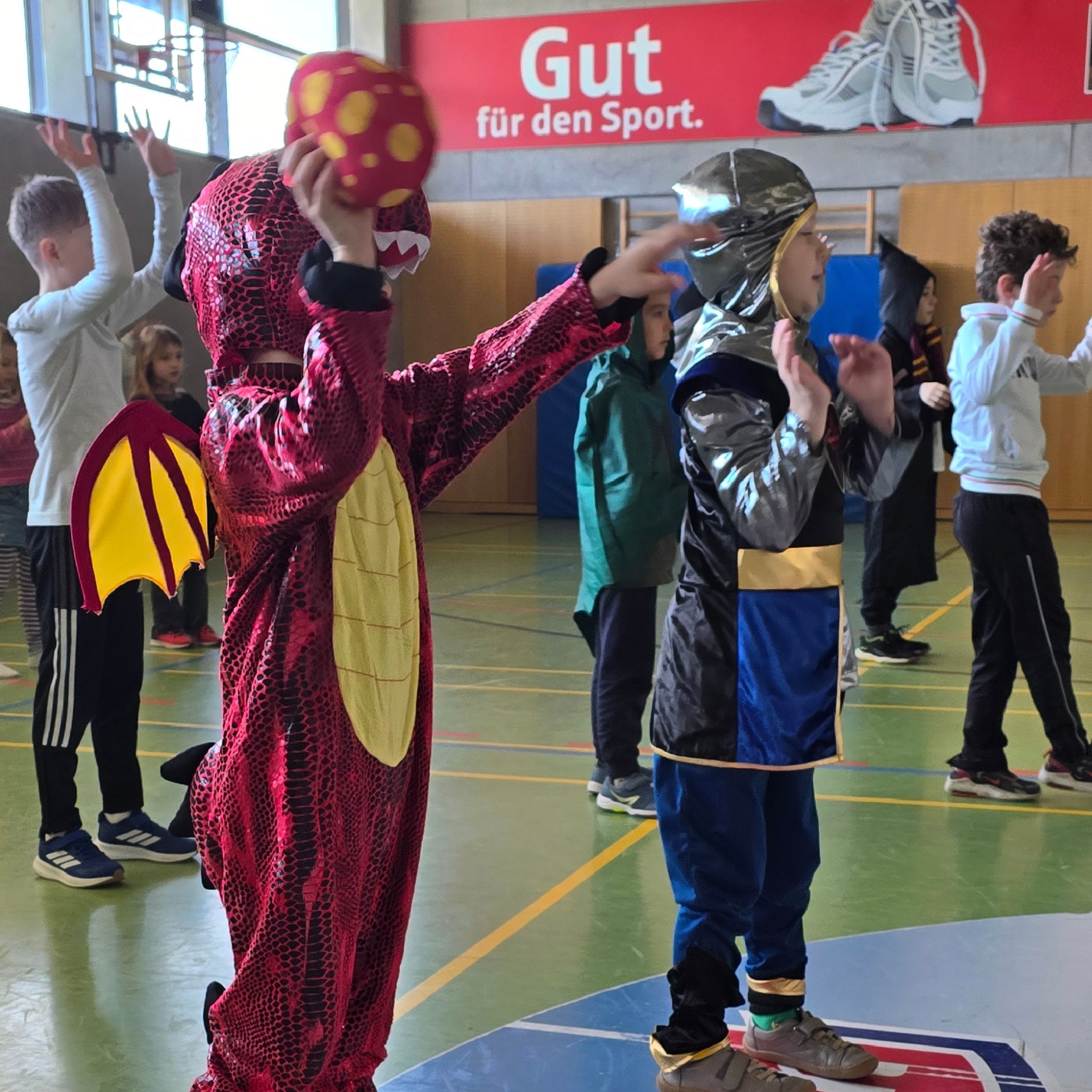 Kinder in fantasievollen Kostümen (Drache mit gepunktetem Ball, Ritter) in einer Turnhalle. Im Hintergrund ein Banner: „Gut für den Sport.“